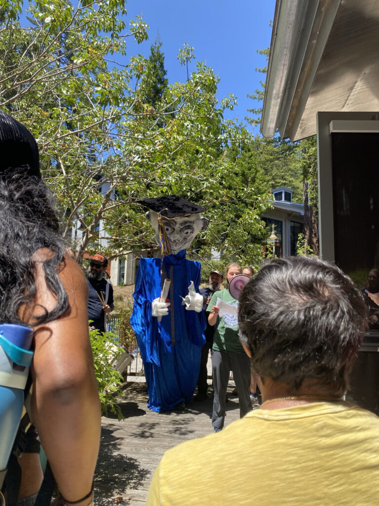 a larger-than-life sized paper mace puppet wearing academic regalia looms behind a young woman at a rally